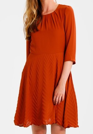 Kobieta w sukience za kolano w kolorze spalonym pomarańczowym, z rękawami do połowy przedramienia i teksturowanym wzorem jodełki na spódnicy.