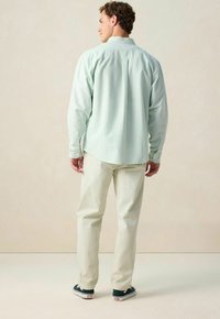 Man in een lichtgroene gestreepte lange mouwen overhemd, met off-white broek en zwarte sneakers, staand en met zijn rug naar de camera gericht tegen een effen achtergrond.