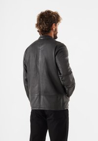 Veste en cuir noir avec une texture lisse, dotée d'un col montant et de coutures discrètes, associée à un pantalon sombre.