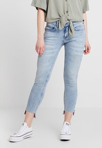 Jeans in denim azzurro chiaro con una vestibilità slim, caratterizzati da dettagli con orlo grezzo alla caviglia. Abbinati a una blusa annodata verde chiaro e a sneakers bianche.