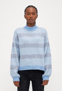 Pull en bleu clair avec un col montant, arborant un motif en bleu foncé et marron. Coupe ample avec des bords-côtes aux poignets et à l'ourlet.