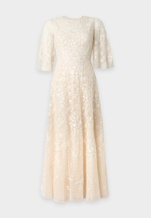 Mujer de pie con un vestido largo y transparente de color crema con bordados florales y mangas cortas acampanadas, combinado con sandalias beige de tacón alto.