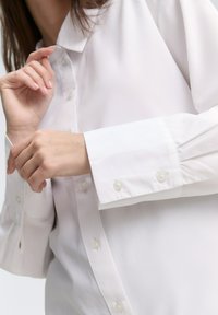 Camicia bianca con colletto a punta, bottoni in perla e maniche lunghe con polsini abbottonati. Tessuto dalla texture liscia.