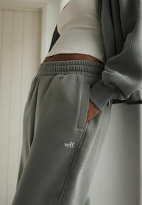 Pantaloni sportivi grigi con vita elastica, tasche laterali e dettaglio ricamato "self." Texture morbida, vestibilità rilassata, abbinati a una maglietta bianca.