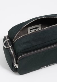 Bolso negro de nailon tipo bandolera con cremalleras plateadas, que cuenta con un diseño de doble compartimento y una etiqueta de logo en el interior. Textura suave y acentos mínimos.