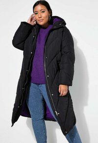 Lång svart puffjacka med lila foder, utrustad med huva, diagonala kviltningar och silverdetaljer. Bärs över en lila tröja och blå jeans.