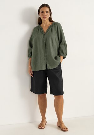 Chemise en lin verte à boutons, avec des manches trois-quarts, assortie à un short noir et des sandales plates beiges, sur fond neutre.