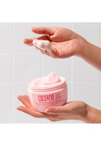 Hårmask i en rosa, rund burk hålls i händer; krämig rosa produkt tas ut; etiketten inkluderar "Coco & Eve Sweet Repair."