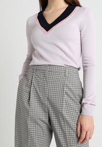 Helllila Pullover mit einem schwarz-pinken V-Ausschnitt, kombiniert mit hochgeschnittenen, karierten Hosen mit Faltendetails.