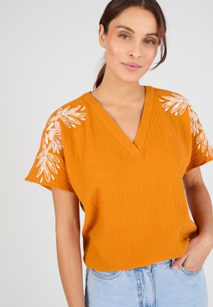 Mujer con blusa naranja texturizada con escote en V y bordado floral blanco en las mangas, combinada con vaqueros azul claro, posando con la mano en el bolsillo.