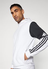 Weißer Hoodie mit schwarzen Ärmeln und drei weißen Streifen am Handgelenk. Der Stoff sieht weich und strukturiert aus, verfügt über einen hohen Kragen und eine Kängurutasche.