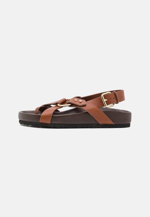 Sandal de couro castanho com sola plana, faixa de tornozelo ajustável e detalhes em metal dourado. Apresenta tiras cruzadas e um design texturizado.