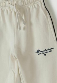 Pantalon de survêtement couleur crème avec ceinture élastique, cordon blanc, poches latérales, passepoil noir, et texte noir indiquant "All round winner Sports Club Italia 1995"