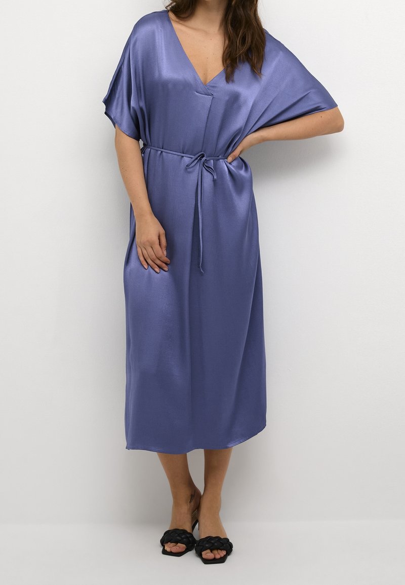 Femme portant une robe midi violette fluide et brillante avec un col en V, des manches courtes et une taille nouée, associée à des sandales slides tressées noires.