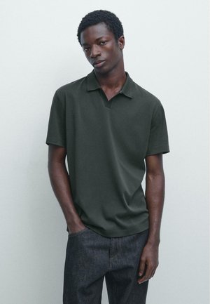 Giovane uomo dalla pelle scura che indossa una polo verde scuro a maniche corte e jeans scuri, in piedi con una mano in tasca davanti a uno sfondo semplice.