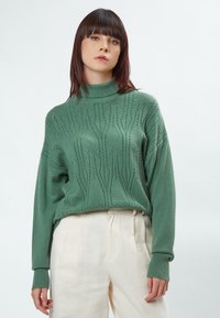 Grüner gestrickter Pullover mit hohem Kragen, der ein strukturiertes Muster, gerippte Bündchen und einen lässigen Schnitt aufweist, kombiniert mit hellen Hosen.