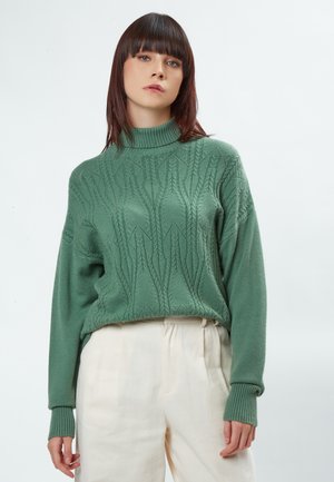 Maglione verde a maglia con collo alto, caratterizzato da un motivo tessuto, polsini a coste e una vestibilità rilassata, abbinato a pantaloni di colore chiaro.