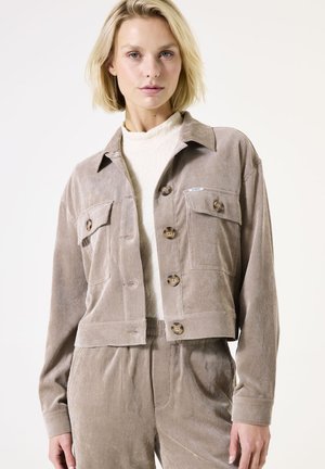 Femme blonde portant une veste beige en velours côtelé boutonnée et un pantalon assorti, sur un haut blanc à col montant, sur un fond clair uni.