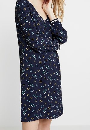 Žena nosící tmavě modré šaty s dlouhým rukávem a malými modrými a žlutými listovými vzory, stojící s jednou rukou ohnutou a rukou u obličeje.