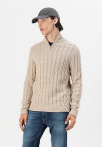 Beige kötött pulóver, kábelmintával, negyed cipzárral, bordázott mandzsettákkal és szegéllyel, kék farmernadrággal és szürke sapkával párosítva.