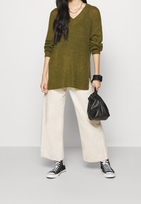 Personne portant un pull à col en V vert olive, un pantalon large beige, des baskets montantes noires, tenant un petit sac à main en cuir noir dans la main droite.