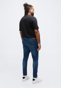 Άνδρας με κοτσίδες στα μαλλιά, φορώντας μαύρο μπλουζάκι, μπλε τζιν και λευκά αθλητικά παπούτσια, στέκεται πλάγια μπροστά σε απλό λευκό φόντο.