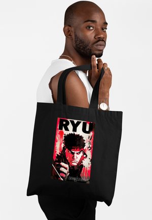 Mężczyzna trzymający czarną torbę typu tote z czerwono-czarną grafiką Ryu z Street Fighter z japońskimi znakami i pogrubionym tekstem.