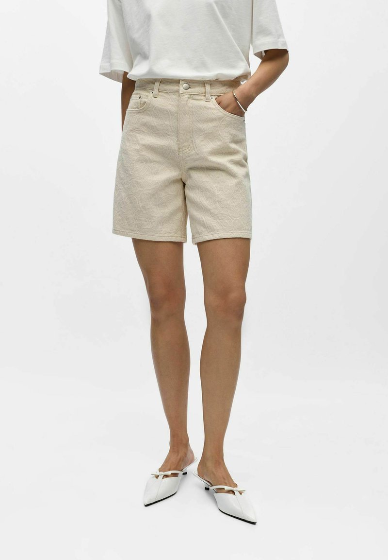 Femme portant un short beige texturé jusqu'aux genoux, une chemise blanche à manches courtes, un bracelet argenté et des ballerines blanches pointues avec un petit nœud.