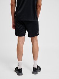 Mand iført sorte atletikshorts, sort skjorte, hvide ankelsokker og sorte sneakers, stående med ryggen mod kameraet på en ensfarvet lys baggrund.