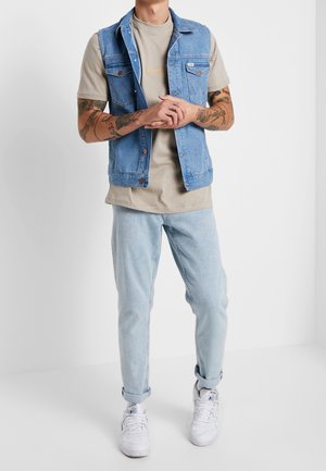 Denimvest med en lys vask, blå, med forlommer og knaplukninger, båret over en beige t-shirt og lyseblå jeans. Hvide sneakers.