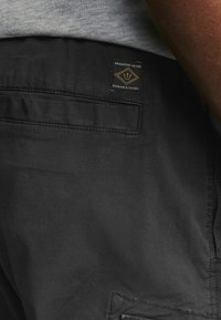 Pantaloni negri cu o buzunar plat ce prezintă un eticheta rectangulară pe care scrie "Produse Durabile de Calitate Premium." Țesătura pare netedă și rezistentă.