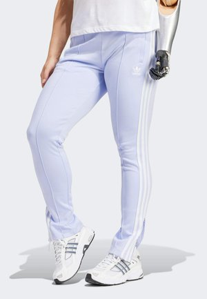 Osoba ubrana w jasnofioletowe spodnie dresowe Adidas z białymi paskami i białe trampki, pokazująca metalową protezę prawej ręki.