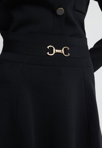 Černé šaty s texturovaným materiálem, širokým pasem a zlatou geometrickou sponou pro zapínání. Přiléhavý střih se strukturovaným designem.