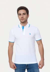 Ung mand i en hvid polo skjorte med blå detaljer, iført jeans, der står op ad en lys baggrund og kigger direkte ind i kameraet.