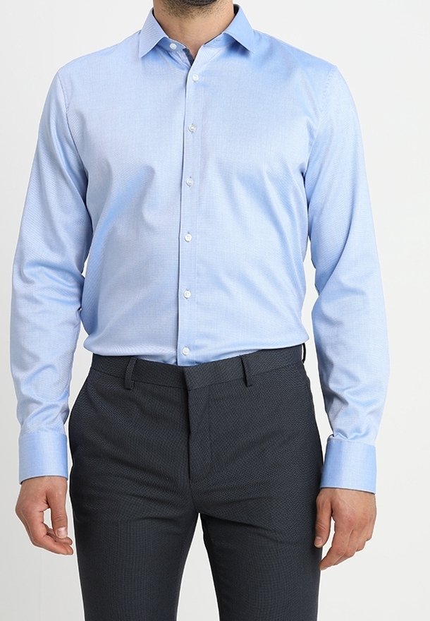 Camicia azzurra chiara con motivo a piccoli quadretti e colletto aperto, abbinata a pantaloni scuri con un motivo. Il tessuto sembra liscio e fresco.