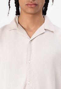 Camicia button-up beige chiaro realizzata in un tessuto morbido e testurizzato, con colletto e pattina frontale con bottoni bianchi.