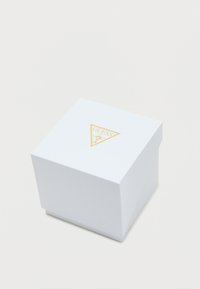 Fehér, textúrázott négyzet alakú ajándékdoboz, arany háromszög logóval a tetején, amelyen a "Guess" márkaneve és díszítő elemek találhatók.