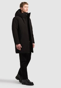 Jeune homme marchant, portant un manteau d'hiver noir à capuche, un pantalon noir et des chaussures noires à lacets sur un fond clair uni.