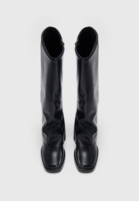 Paire de bottes en cuir noir montant jusqu'aux genoux avec des bouts carrés, debout droite sur un fond blanc uni.