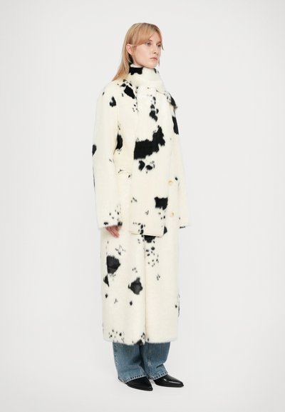 Abrigo largo de lana con un estampado de vaca en blanco y negro, con un cuello alto y dos botones visibles. Combinado con unos vaqueros de denim azul de pierna ancha.