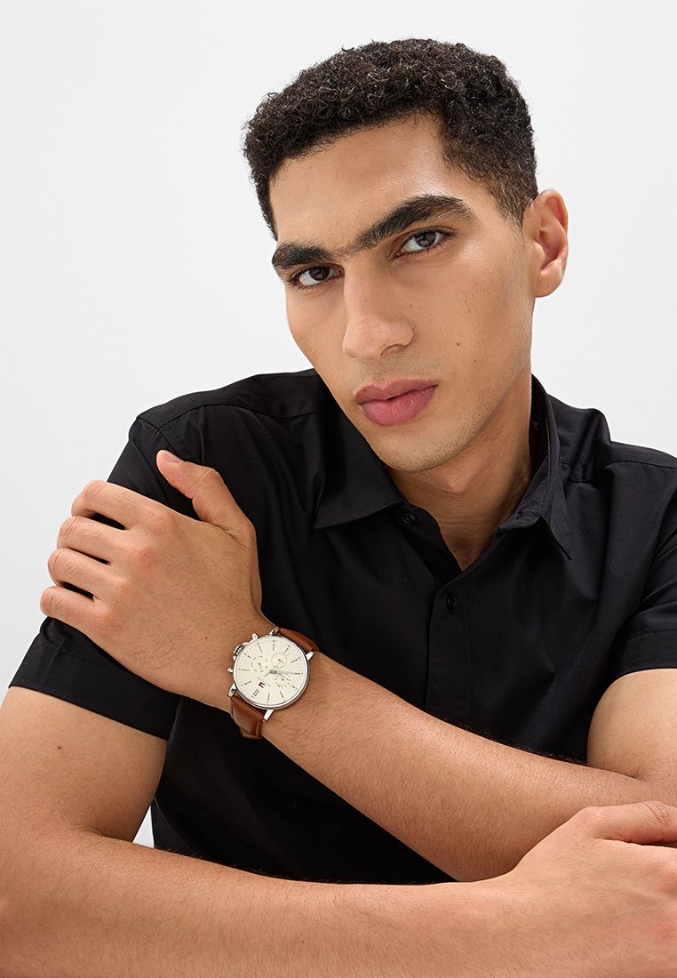 Braunlederarmbanduhr mit einem cremefarbenen Zifferblatt, silbernen Stundenmarkierungen und drei Subdials. Getragen an einem nackten Handgelenk, kombiniert mit einem schwarzen Hemd.