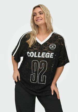 Uśmiechnięta kobieta o długich blond włosach ma na sobie czarną sportową koszulkę z koronki z białym napisem "COLLEGE 92" i krótkimi rękawami w paski, pozując z rękami na biodrach.