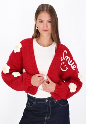 Rode gebreide cardigan met witte bloemenapplicaties op de mouwen en de tekst "Smile". Geribbelde boorden en ontwerp met open voorkant. Witte binnen-top zichtbaar.