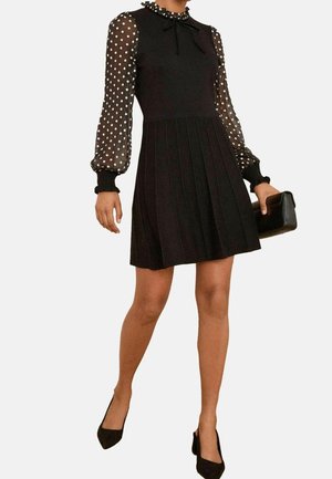 Vrouw draagt een zwarte geplooide jurk met doorschijnende lange mouwen met stippen, houdt een zwarte clutch vast en zwarte gesloten pumps.