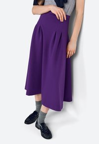 Lila midi szoknya redőkkel, sima textúrával, sötét cipőkkel és szürke zoknikkal párosítva. A modell fehér háttér előtt pózol.