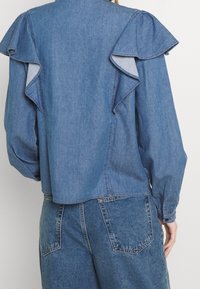 Jeansbluse mit Raffungen auf der Rückseite, langen Ärmeln und einem lockeren Schnitt. Hellblauer Stoff mit einer glatten Textur.