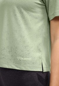 Jasnozielona koszulka sportowa z delikatną teksturą, z bocznym rozcięciem i nadrukowanym logo na dole. Połączona z ciemnoszarymi spodniami.