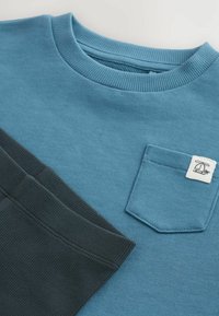Gros plan d'un sweat-shirt bleu avec une petite poche avant et une étiquette blanche, associé à un pantalon en tissu côtelé gris foncé.