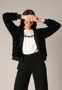 Veste bomber noire en sequins avec un tissu texturé, des poignets en bord-côte et une taille en bord-côte. Portée sur une chemise blanche avec des lettres noires. Pantalons noirs.