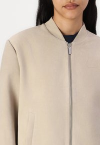 Heledbeez bomber-jakk, mis on valmistatud nahast, funktsioneeriva lõuaga, esiosa tõmblukuga ja küljetaskuga. Sile tekstuur ja puhtad jooned.